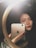 A person is taking a selfie using a smartphone in front of a circular mirror. The phone has a prominent logo on its back. The lighting is dim, casting shadows, and the focus is on the person's face and hand holding the phone.