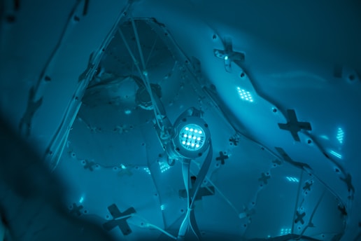 A futuristic and abstract interior environment with complex geometric structures and illuminated elements. The setting is predominantly blue with a soft, glowing light emanating from various points, creating a serene yet mysterious atmosphere.