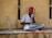 A man with a red turban is seated cross-legged against a textured beige wall, playing a stringed musical instrument. Another decorated, stick-like instrument is placed beside him. He wears a white shirt and blue jeans, and appears focused on playing music. The scene conveys a sense of tradition and culture.