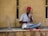 A man with a red turban is seated cross-legged against a textured beige wall, playing a stringed musical instrument. Another decorated, stick-like instrument is placed beside him. He wears a white shirt and blue jeans, and appears focused on playing music. The scene conveys a sense of tradition and culture.