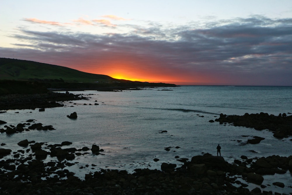 person standing on rocks surrounded by body of water overlooking sunset