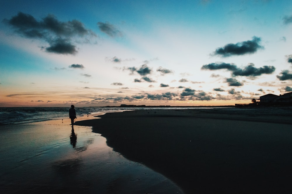 Silhouette einer Person, die neben dem Ozean unter blauem und bewölktem Himmel während des Sonnenuntergangs spazieren geht