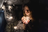 A person holding a string of glowing lights, creating a warm and cozy atmosphere. The surroundings are dimly lit with additional lights draped around, casting soft shadows.