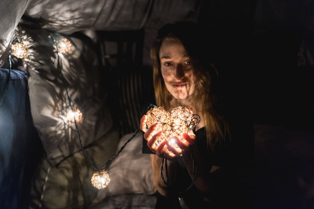 A person holding a string of glowing lights, creating a warm and cozy atmosphere. The surroundings are dimly lit with additional lights draped around, casting soft shadows.