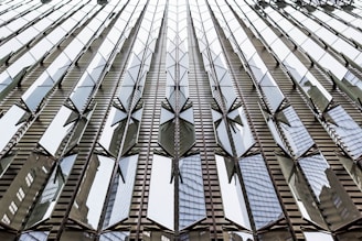 A modern building facade with reflective glass panels arranged in a geometric pattern. The reflection of neighboring structures is visible on the glass, creating an abstract and dynamic visual effect.
