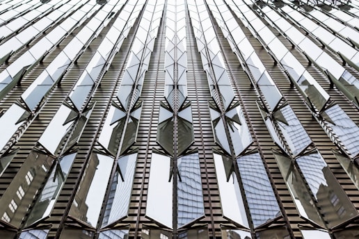 A modern building facade with reflective glass panels arranged in a geometric pattern. The reflection of neighboring structures is visible on the glass, creating an abstract and dynamic visual effect.