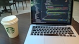 A laptop displaying code is placed on a round wooden table next to a disposable coffee cup with a Starbucks logo. The background features several patio-style chairs and tables on a light-tiled floor.