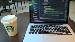 A laptop displaying code is placed on a round wooden table next to a disposable coffee cup with a Starbucks logo. The background features several patio-style chairs and tables on a light-tiled floor.