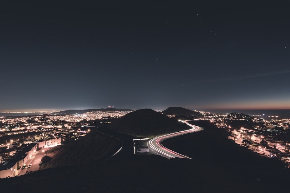 Foto de lapso de tiempo de coches que corren por la carretera con curvas al lado de la montaña rodeados de edificios y establecimientos por la noche