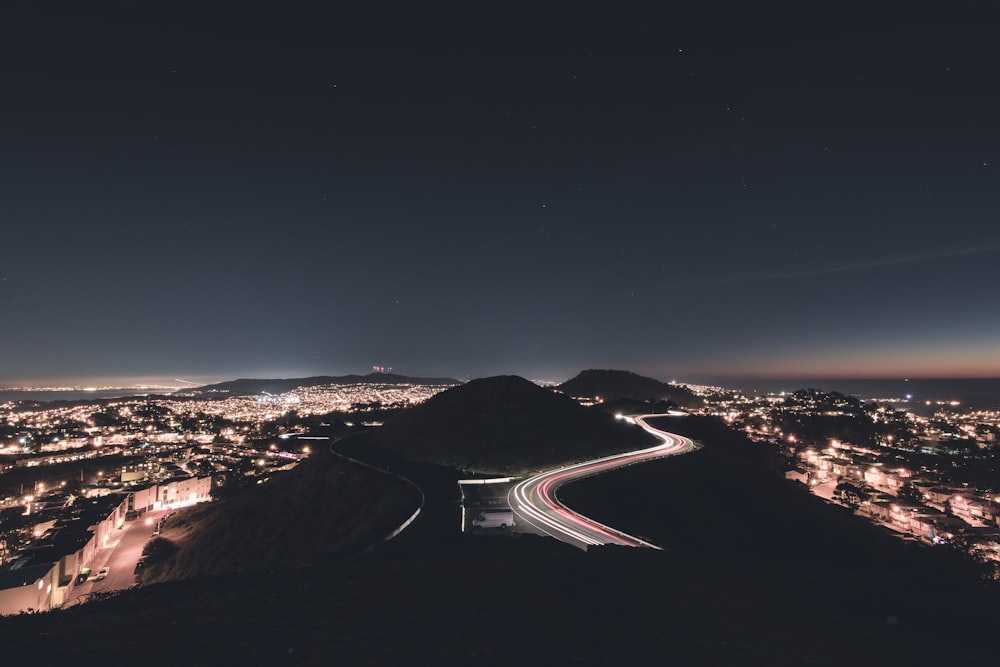 Foto de lapso de tempo de carros rodando em estrada curvilínea ao lado da montanha cercada por edifícios e estabelecimentos em Nighr