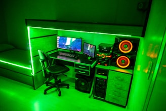 A modern gaming setup featuring a desk with a computer monitor displaying a space-themed screensaver, accompanied by a keyboard and mouse. The desk is integrated with LED strip lighting that casts a vibrant green hue throughout the room. Adjacent to the computer setup is a powerful sound system with large speakers highlighted by orange lighting. A comfortable swivel chair is positioned in front of the desk, and a bed with matching LED lighting is visible in the background.