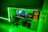 A modern gaming setup featuring a desk with a computer monitor displaying a space-themed screensaver, accompanied by a keyboard and mouse. The desk is integrated with LED strip lighting that casts a vibrant green hue throughout the room. Adjacent to the computer setup is a powerful sound system with large speakers highlighted by orange lighting. A comfortable swivel chair is positioned in front of the desk, and a bed with matching LED lighting is visible in the background.