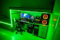 A modern gaming setup featuring a desk with a computer monitor displaying a space-themed screensaver, accompanied by a keyboard and mouse. The desk is integrated with LED strip lighting that casts a vibrant green hue throughout the room. Adjacent to the computer setup is a powerful sound system with large speakers highlighted by orange lighting. A comfortable swivel chair is positioned in front of the desk, and a bed with matching LED lighting is visible in the background.