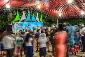 A vibrant nighttime gathering under a large canopy structure with colorful geometric decorations. Blurred figures suggest motion and activity, creating a dynamic atmosphere. The stage features a backdrop sign with text and musical notes, and bright lighting enhances the lively ambiance.