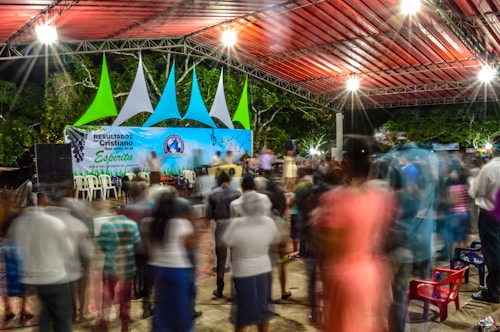 A vibrant nighttime gathering under a large canopy structure with colorful geometric decorations. Blurred figures suggest motion and activity, creating a dynamic atmosphere. The stage features a backdrop sign with text and musical notes, and bright lighting enhances the lively ambiance.