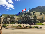 A large, elegant building with green and beige architecture, resembling a hotel or historical site, stands against a backdrop of mountains and a clear blue sky. Three flags are displayed prominently in the foreground, including the Canadian flag. The area is landscaped with greenery and flowering plants, and there are several people walking around.