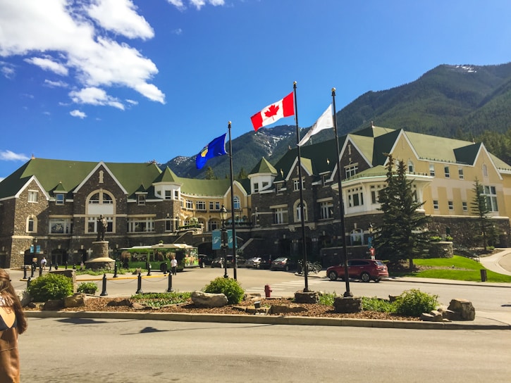 A large, elegant building with green and beige architecture, resembling a hotel or historical site, stands against a backdrop of mountains and a clear blue sky. Three flags are displayed prominently in the foreground, including the Canadian flag. The area is landscaped with greenery and flowering plants, and there are several people walking around.