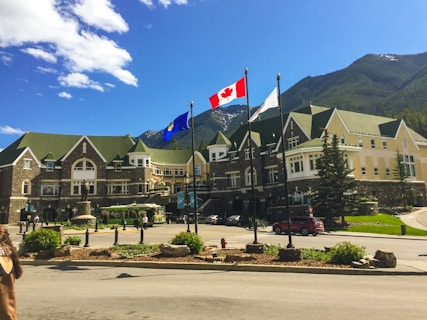 A large, elegant building with green and beige architecture, resembling a hotel or historical site, stands against a backdrop of mountains and a clear blue sky. Three flags are displayed prominently in the foreground, including the Canadian flag. The area is landscaped with greenery and flowering plants, and there are several people walking around.