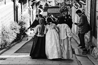 A group of women wearing traditional clothing are walking down a narrow street adorned with a variety of architectural and natural elements. The scene includes buildings with distinct design patterns on both sides, and a man on the right side of the image appears to be engaged with a mobile device. Other people are visible in the background, adding a sense of liveliness to the setting.