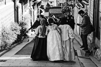 A group of women wearing traditional clothing are walking down a narrow street adorned with a variety of architectural and natural elements. The scene includes buildings with distinct design patterns on both sides, and a man on the right side of the image appears to be engaged with a mobile device. Other people are visible in the background, adding a sense of liveliness to the setting.