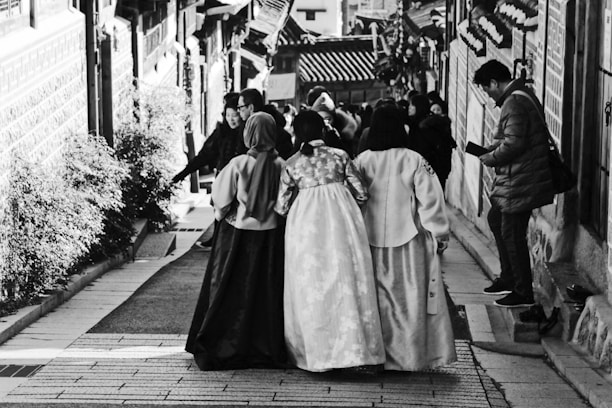 A group of women wearing traditional clothing are walking down a narrow street adorned with a variety of architectural and natural elements. The scene includes buildings with distinct design patterns on both sides, and a man on the right side of the image appears to be engaged with a mobile device. Other people are visible in the background, adding a sense of liveliness to the setting.