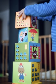 A child wearing a blue striped sleeve is stacking colorful, illustrated blocks. Each block has a different design, including animals, numbers, and vehicles. The blocks are arranged vertically, and the child's hand is placing a block on top of the stack.