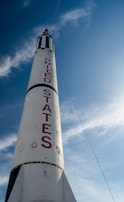 A towering white rocket with bold red letters spelling 'United States' against a bright blue sky. The rocket has a pointed tip and is partially adorned with black stripes.