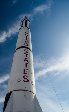 A towering white rocket with bold red letters spelling 'United States' against a bright blue sky. The rocket has a pointed tip and is partially adorned with black stripes.