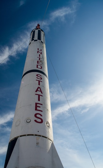 A towering white rocket with bold red letters spelling 'United States' against a bright blue sky. The rocket has a pointed tip and is partially adorned with black stripes.