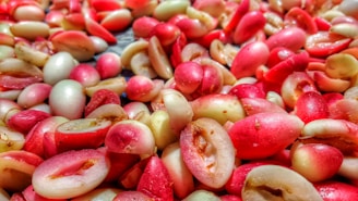 A close-up view of numerous small, pink and light cream-colored fruits piled together. Their shiny surfaces suggest freshness, with some fruits split open, revealing the seed inside.