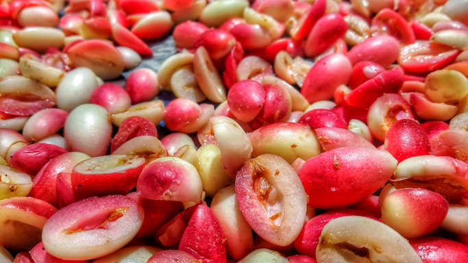 A close-up view of numerous small, pink and light cream-colored fruits piled together. Their shiny surfaces suggest freshness, with some fruits split open, revealing the seed inside.
