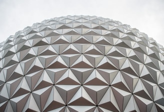A detailed view of a large geodesic dome with an intricate pattern of triangular panels. The panels are arranged symmetrically, creating a sense of geometric precision and architectural innovation. The overcast sky provides a neutral backdrop, emphasizing the design of the structure.