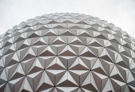 A detailed view of a large geodesic dome with an intricate pattern of triangular panels. The panels are arranged symmetrically, creating a sense of geometric precision and architectural innovation. The overcast sky provides a neutral backdrop, emphasizing the design of the structure.