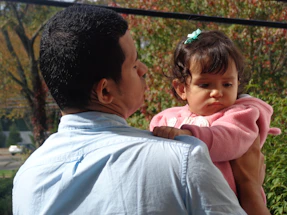A person is holding a young child, engaging in a tender and protective manner. The child is wearing a pink sweater and has a small bow in their hair, looking slightly pensive. The background is filled with greenery and hints of autumn foliage, suggesting an outdoor setting.
