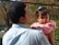 A person is holding a young child, engaging in a tender and protective manner. The child is wearing a pink sweater and has a small bow in their hair, looking slightly pensive. The background is filled with greenery and hints of autumn foliage, suggesting an outdoor setting.