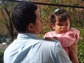 A person is holding a young child, engaging in a tender and protective manner. The child is wearing a pink sweater and has a small bow in their hair, looking slightly pensive. The background is filled with greenery and hints of autumn foliage, suggesting an outdoor setting.