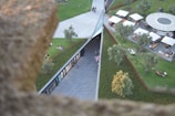 Aerial view of a landscaped public space with grassy areas, trees, and pathways. People are sitting and relaxing on the grass, while others are seated at tables under large umbrellas. A modern structure with glass windows runs through the center, surrounded by greenery.