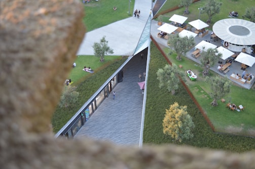 Aerial view of a landscaped public space with grassy areas, trees, and pathways. People are sitting and relaxing on the grass, while others are seated at tables under large umbrellas. A modern structure with glass windows runs through the center, surrounded by greenery.
