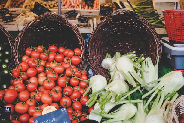 A vibrant market display featuring woven baskets full of fresh produce. Bright red tomatoes spill out of one basket, while another contains crisp white fennel bulbs. The scene is set against a backdrop of various vegetables, including carrots and greens.