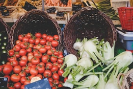 A vibrant market display featuring woven baskets full of fresh produce. Bright red tomatoes spill out of one basket, while another contains crisp white fennel bulbs. The scene is set against a backdrop of various vegetables, including carrots and greens.