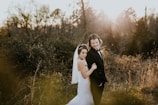 A bride and groom are standing together in a natural, wooded setting. The bride is wearing a white wedding dress with a veil, while the groom is dressed in a black tuxedo with a bow tie. The sunlight creates a warm, glowing effect around them, highlighting the romantic atmosphere.
