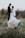 A bouquet of white and peach flowers is prominently placed in the foreground on a natural grassy setting, while in the background, a slightly out-of-focus wedding couple gazes at each other, creating a serene and romantic atmosphere.