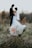 A bouquet of white and peach flowers is prominently placed in the foreground on a natural grassy setting, while in the background, a slightly out-of-focus wedding couple gazes at each other, creating a serene and romantic atmosphere.