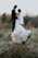 A bouquet of white and peach flowers is prominently placed in the foreground on a natural grassy setting, while in the background, a slightly out-of-focus wedding couple gazes at each other, creating a serene and romantic atmosphere.