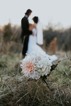A bouquet of white and peach flowers is prominently placed in the foreground on a natural grassy setting, while in the background, a slightly out-of-focus wedding couple gazes at each other, creating a serene and romantic atmosphere.