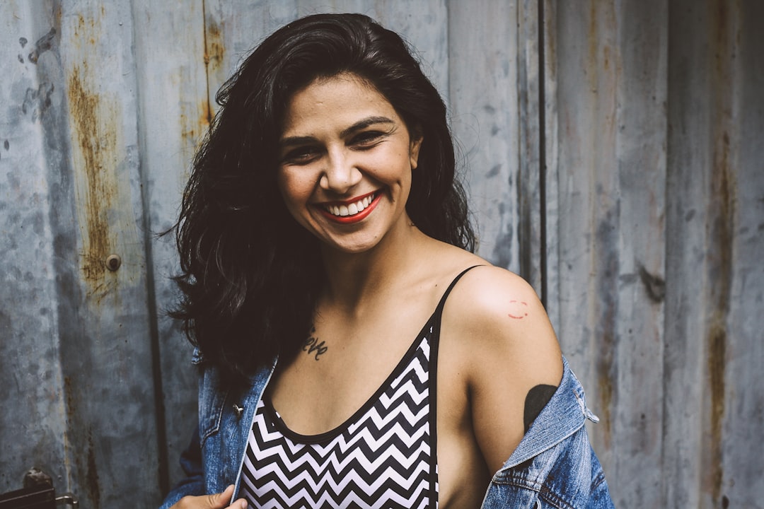 woman wearing white and black chevron print spaghetti-strap top with blue denim jacket standing behind gray wall photography