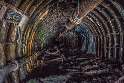 A rusty and abandoned underground tunnel with visible pipes and structural supports. The walls and ceiling are covered in dark and mossy textures, suggesting decay and age. The lighting is dim, creating a mysterious and eerie atmosphere.