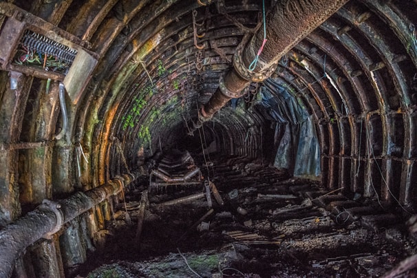 A rusty and abandoned underground tunnel with visible pipes and structural supports. The walls and ceiling are covered in dark and mossy textures, suggesting decay and age. The lighting is dim, creating a mysterious and eerie atmosphere.