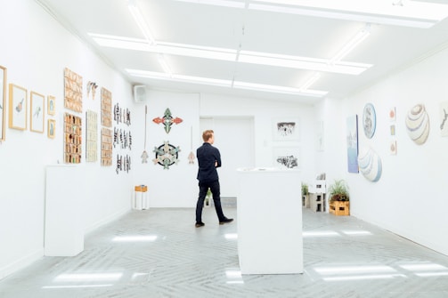 A person stands in a modern art gallery, surrounded by artwork hung on white walls. The gallery features various art pieces including paintings and sculptures. The lighting is bright and evenly distributed, creating a clean and open atmosphere. Plants add a touch of greenery to the otherwise minimalistic space.