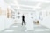 A person stands in a modern art gallery, surrounded by artwork hung on white walls. The gallery features various art pieces including paintings and sculptures. The lighting is bright and evenly distributed, creating a clean and open atmosphere. Plants add a touch of greenery to the otherwise minimalistic space.